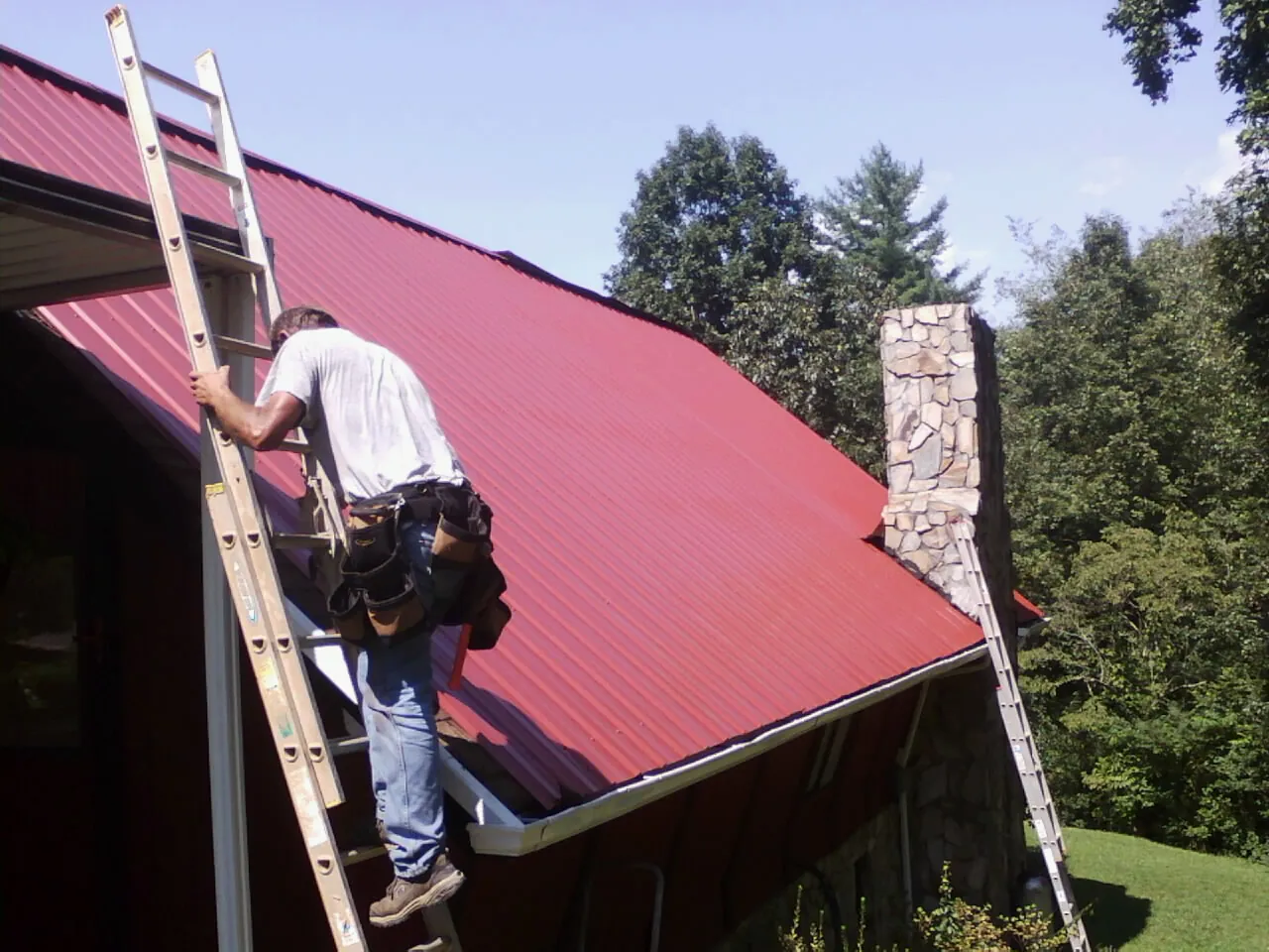 Roofer on a ladder beside a red metal roof and stone chimney — Soffit & Fascia Repair by Roofing Brothers in Whitehall